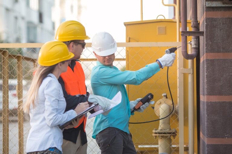 Three workers conducting a site inspection near industrial gas piping, using detection equipment and checklists—illustrating the use of a job hazard analysis checklist to identify potential risks.