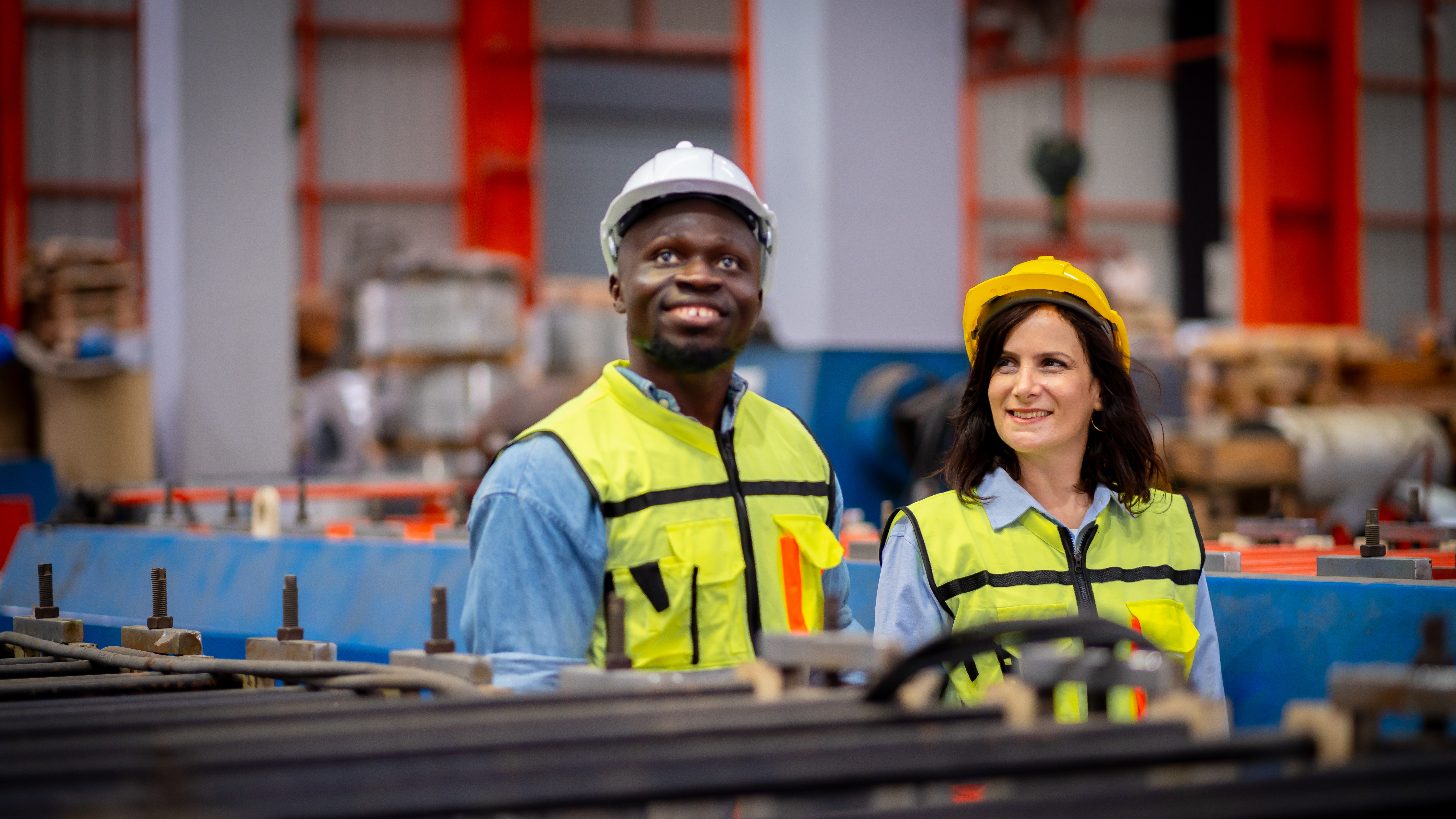A male and female manufacturing engineer smiling with hard hats, goggles, and high visibility vests. An example of following a manufacturing risk assessment checklist on the job.