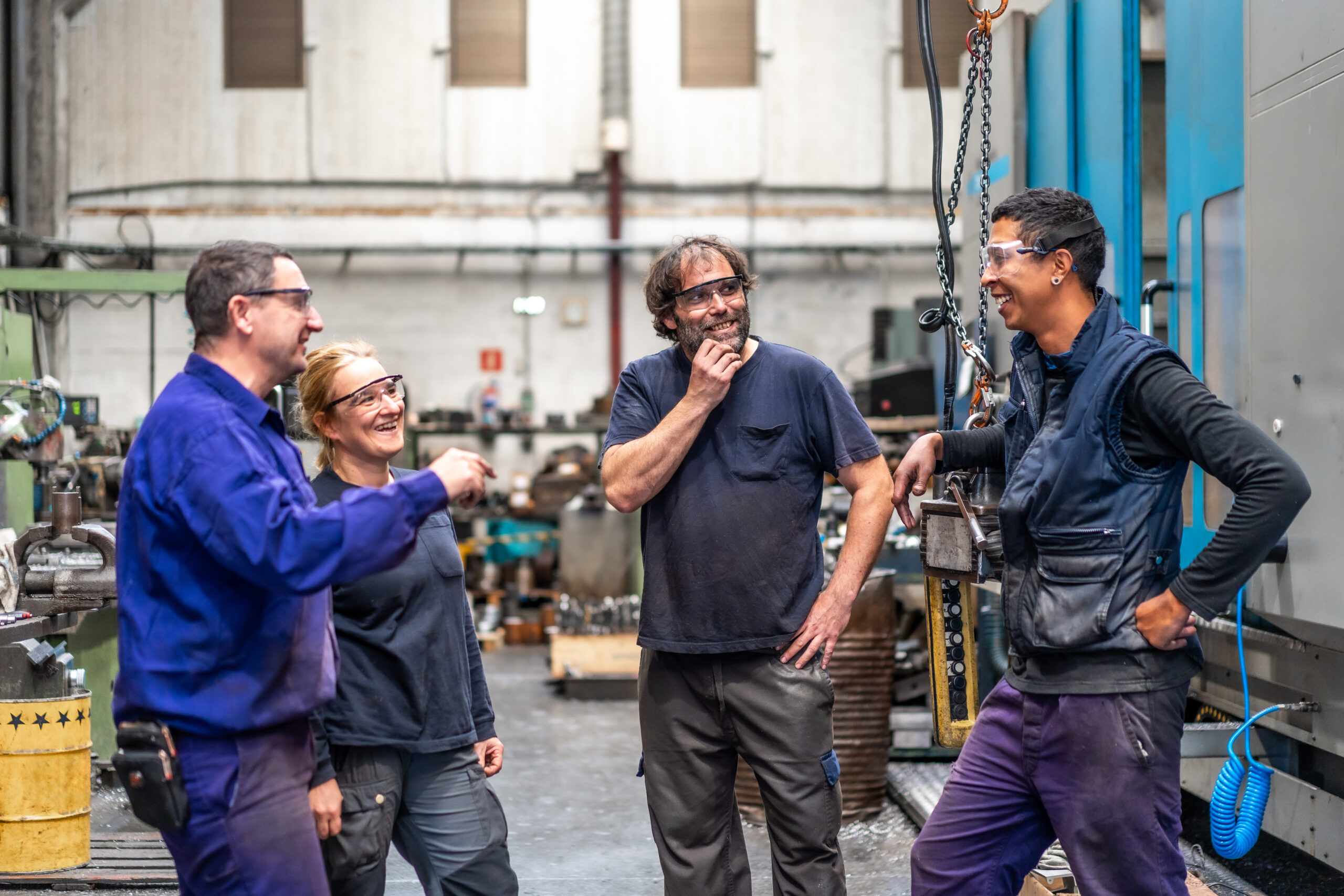 Group of manufacturing workers wearing safety glasses gather around machinery, sharing a lighthearted moment on the shop floor. The team appears engaged and collaborative, reflecting manufacturing best practices that promote strong communication, safety awareness, and positive workplace culture in an industrial environment