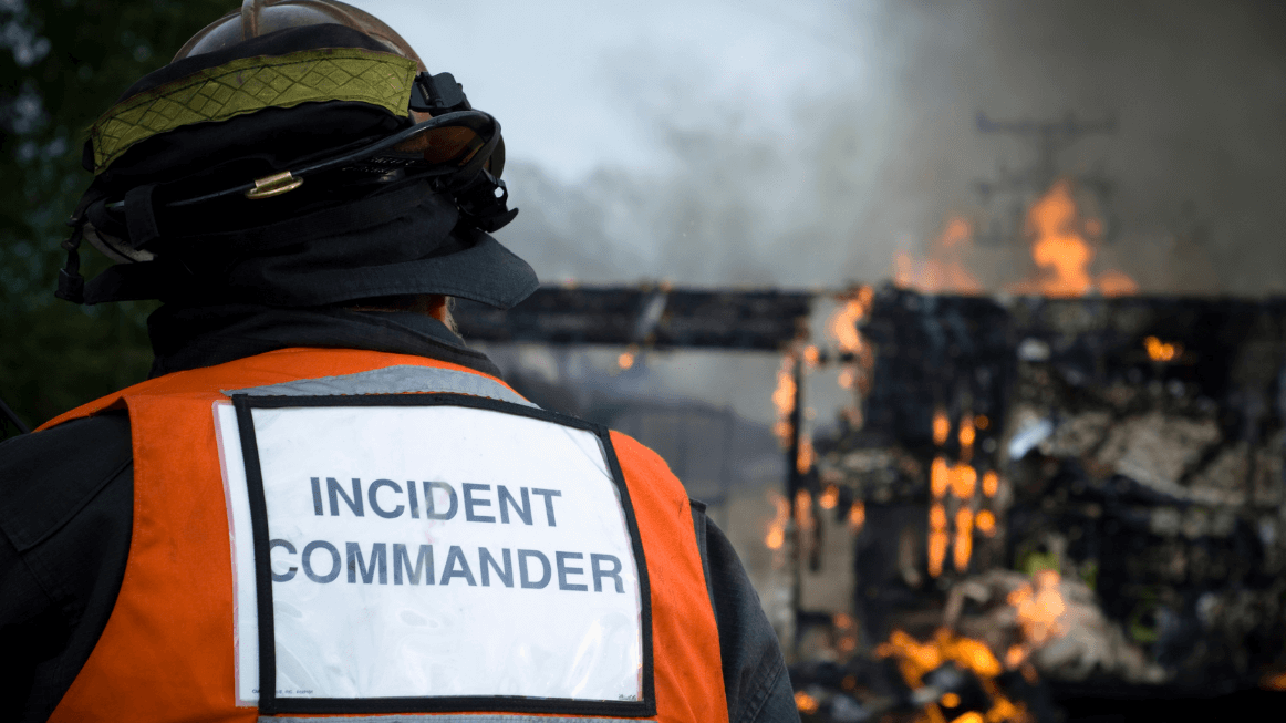 Incident Commander in safety gear standing in front of a burning structure, representing emergency response and root cause investigation in high-risk environments.