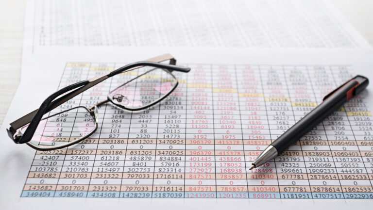 A close-up image of spreadsheets in EHS management, showing rows of multicolored data with a black pen and eyeglasses resting on the paper.
