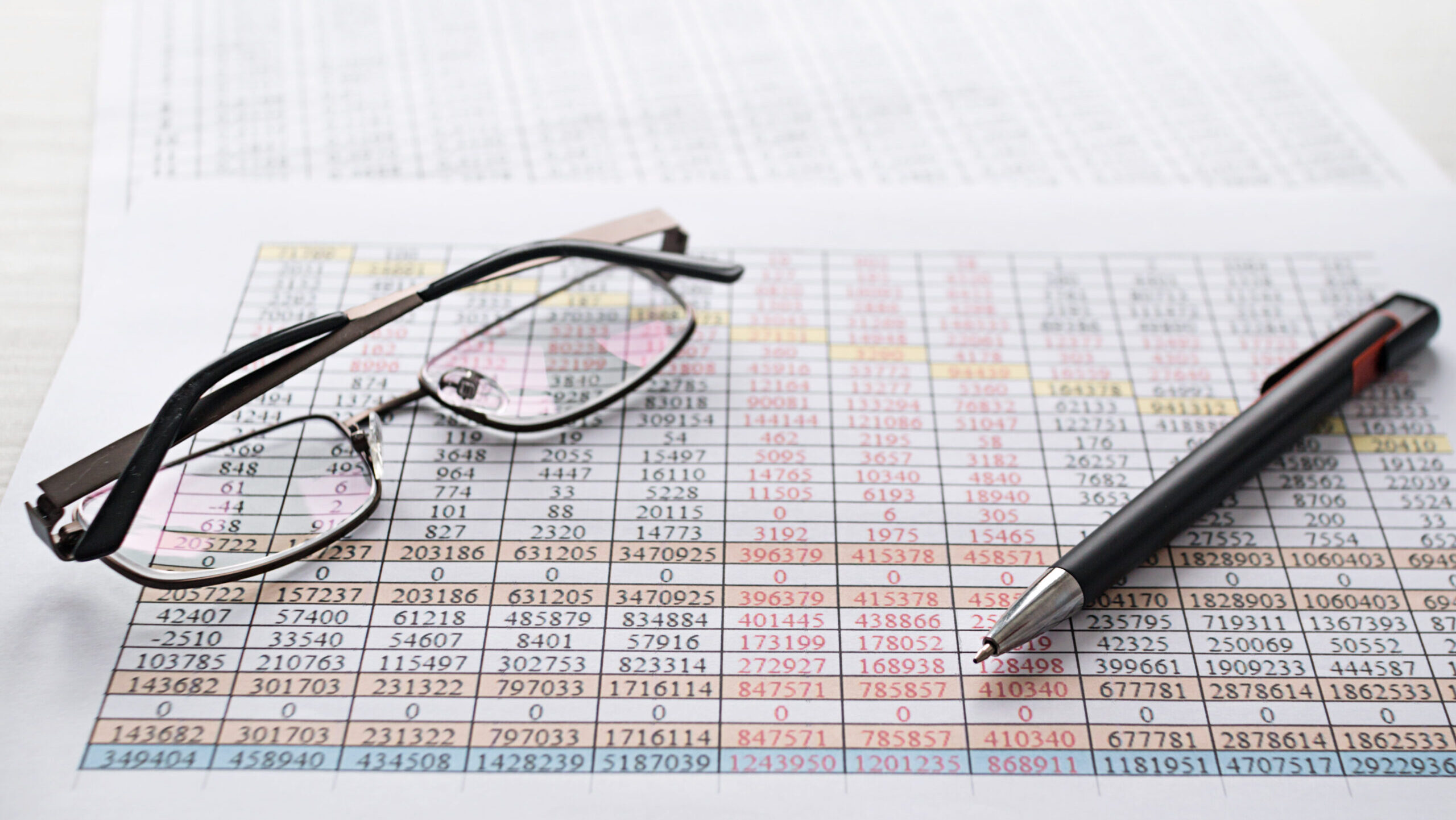 A close-up image of spreadsheets in EHS management, showing rows of multicolored data with a black pen and eyeglasses resting on the paper.