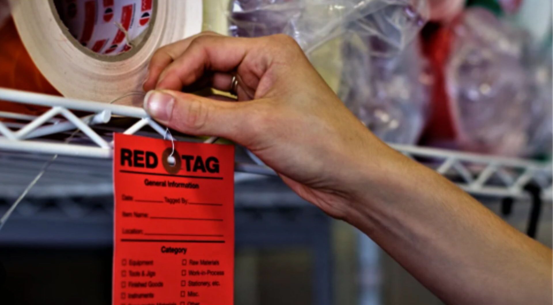 A hand attaches a red tag to a wire shelf as part of a 5S red tag initiative, used to identify items for evaluation, relocation, or disposal in workplace organization efforts.