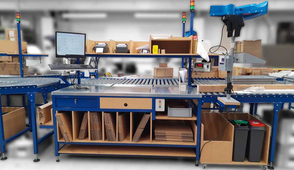 A pack table in front of a conveyor belt with boxes down below, a computer screen to the left side, and andon lights above to the right. Demonstrates how andon systems are integrated with the setup of a work area so they're easy to see from anywhere on the production floor.