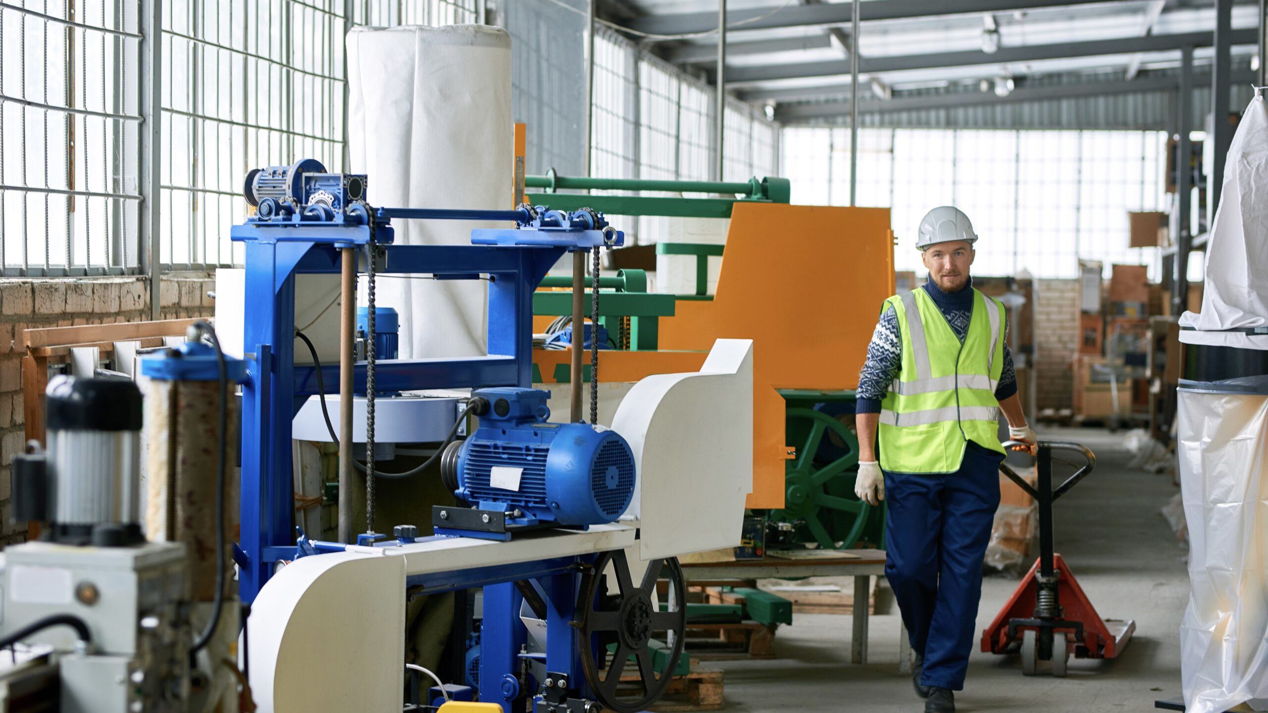 Worker in high-visibility vest and hard hat pulling a pallet jack in an industrial setting, surrounded by machinery, illustrating the importance of following factory safety rules