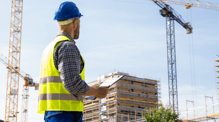 A construction site supervisor in a safety vest and hard hat holds a clipboard while observing active cranes and scaffolding, representing actions taken during an OSHA inspection