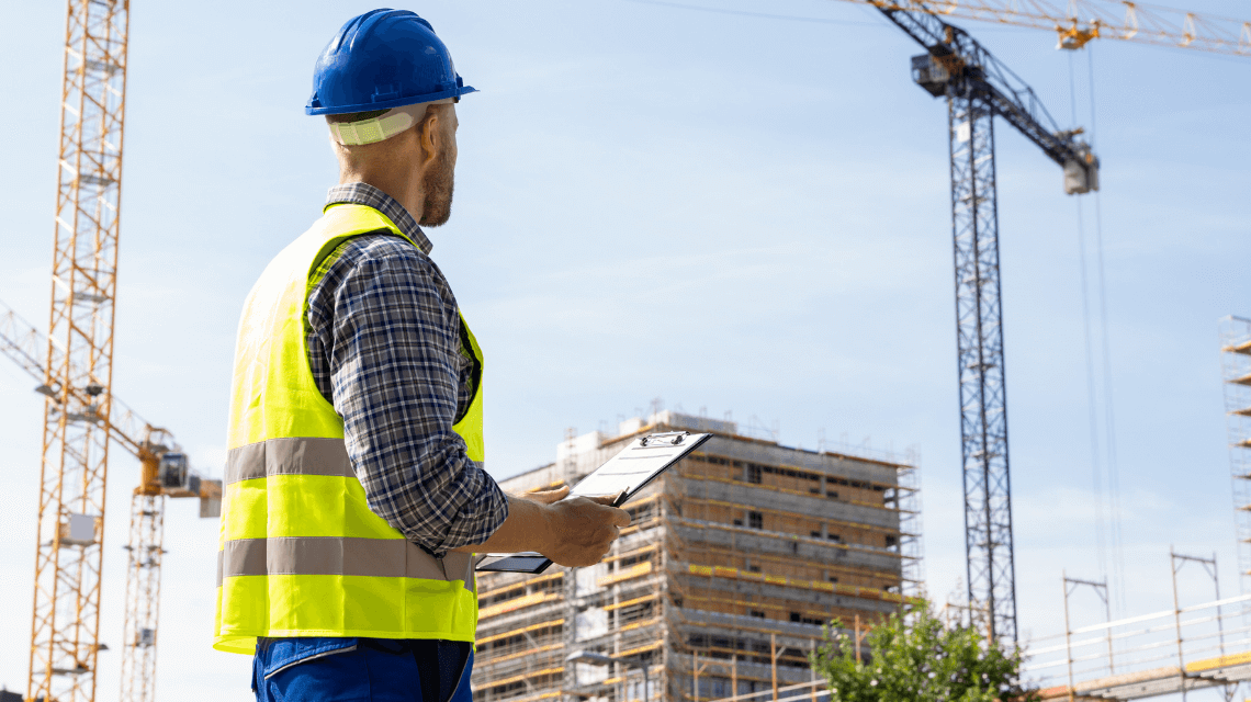 A construction site supervisor in a safety vest and hard hat holds a clipboard while observing active cranes and scaffolding, representing actions taken during an OSHA inspection