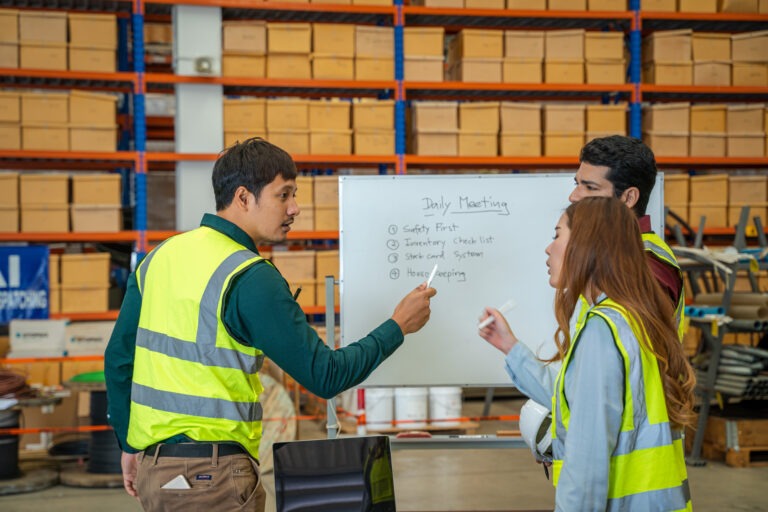 Three warehouse employees in high-visibility vests gather around a whiteboard during a daily meeting in a distribution center. The board lists discussion points including safety, inventory checklists, and housekeeping. This scene illustrates best practices for onboarding by promoting team communication, safety awareness, and operational procedures from day one.
