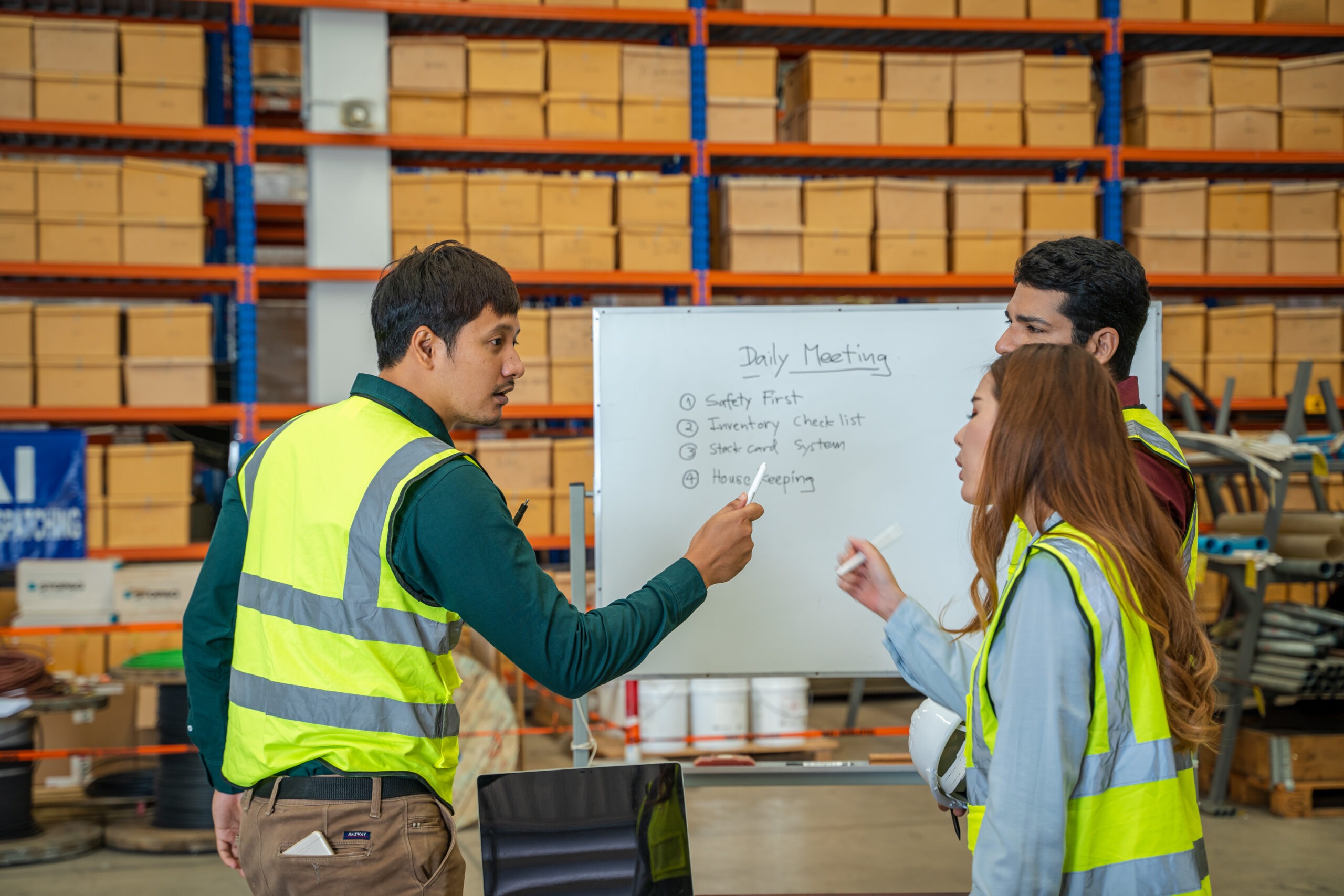 Three warehouse employees in high-visibility vests gather around a whiteboard during a daily meeting in a distribution center. The board lists discussion points including safety, inventory checklists, and housekeeping. This scene illustrates best practices for onboarding by promoting team communication, safety awareness, and operational procedures from day one.