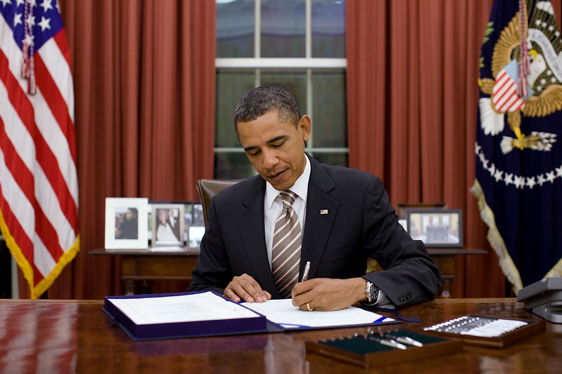 US President Barack Obama signing the Food Safety Modernization Act in the Oval Office. He is sitting at a desk with the American flag on his right and the Presidential flag on his left.