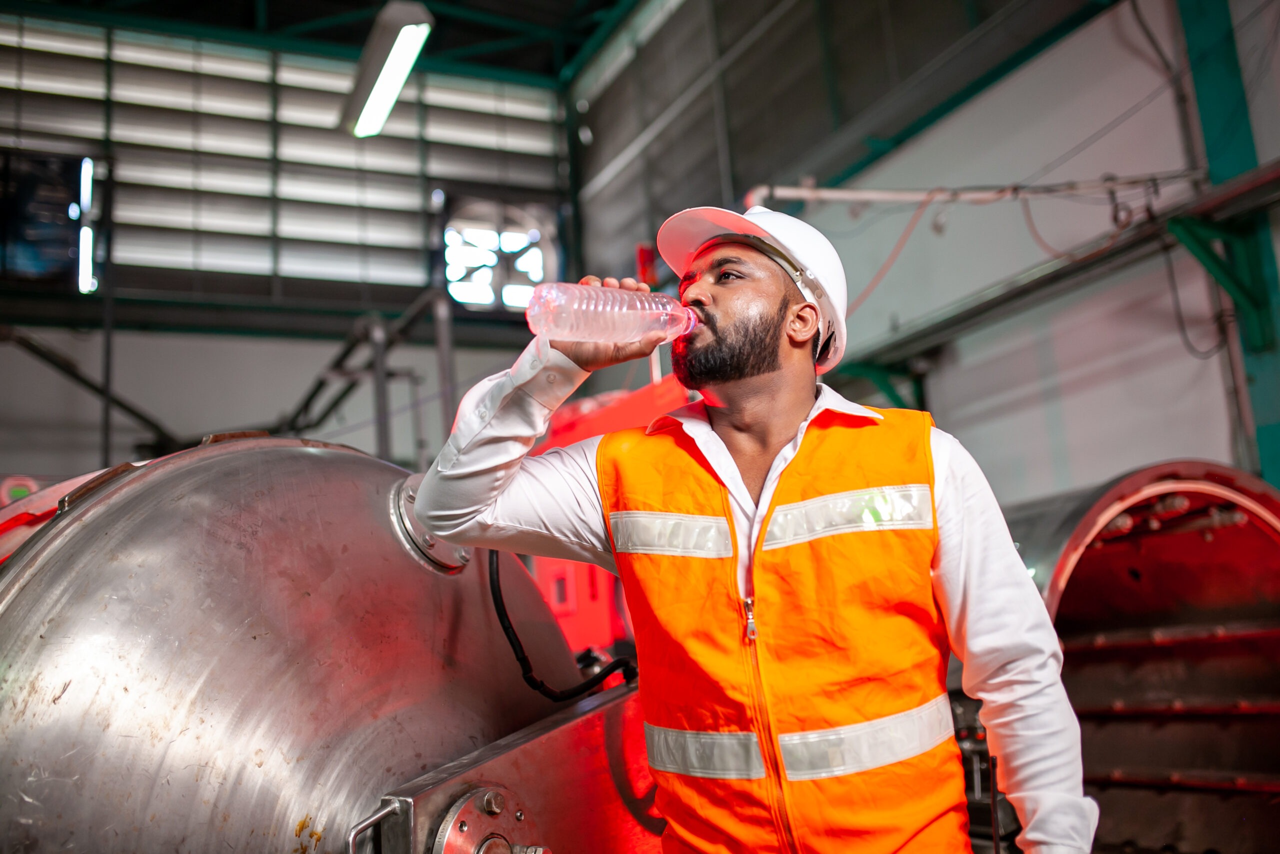 Factory worker in a hard hat and reflective vest drinking water to stay cool, demonstrating summer safety tips for preventing heat illness on the job.