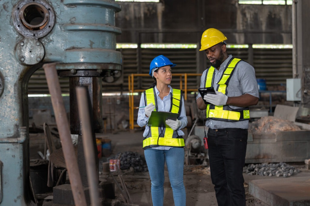 Two engineers wearing hard hats and high-visibility vests discussing EHS performance metrics inside an industrial facility, using a clipboard and tablet to review workplace safety data.