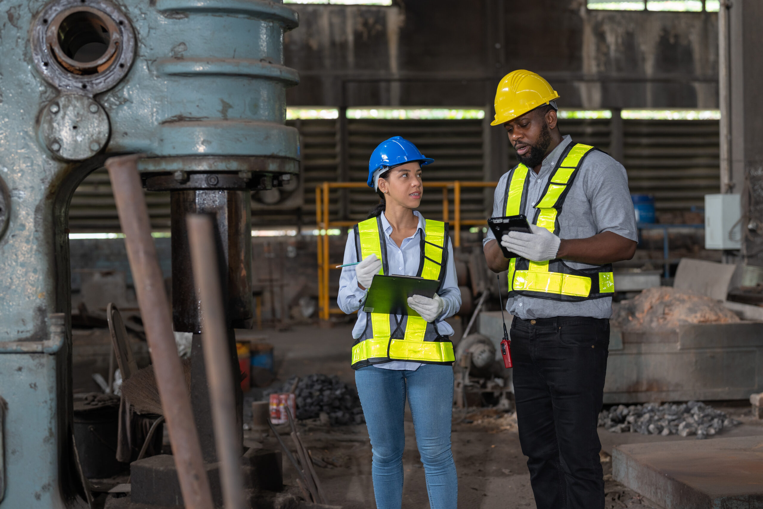 Two engineers wearing hard hats and high-visibility vests discussing EHS performance metrics inside an industrial facility, using a clipboard and tablet to review workplace safety data.