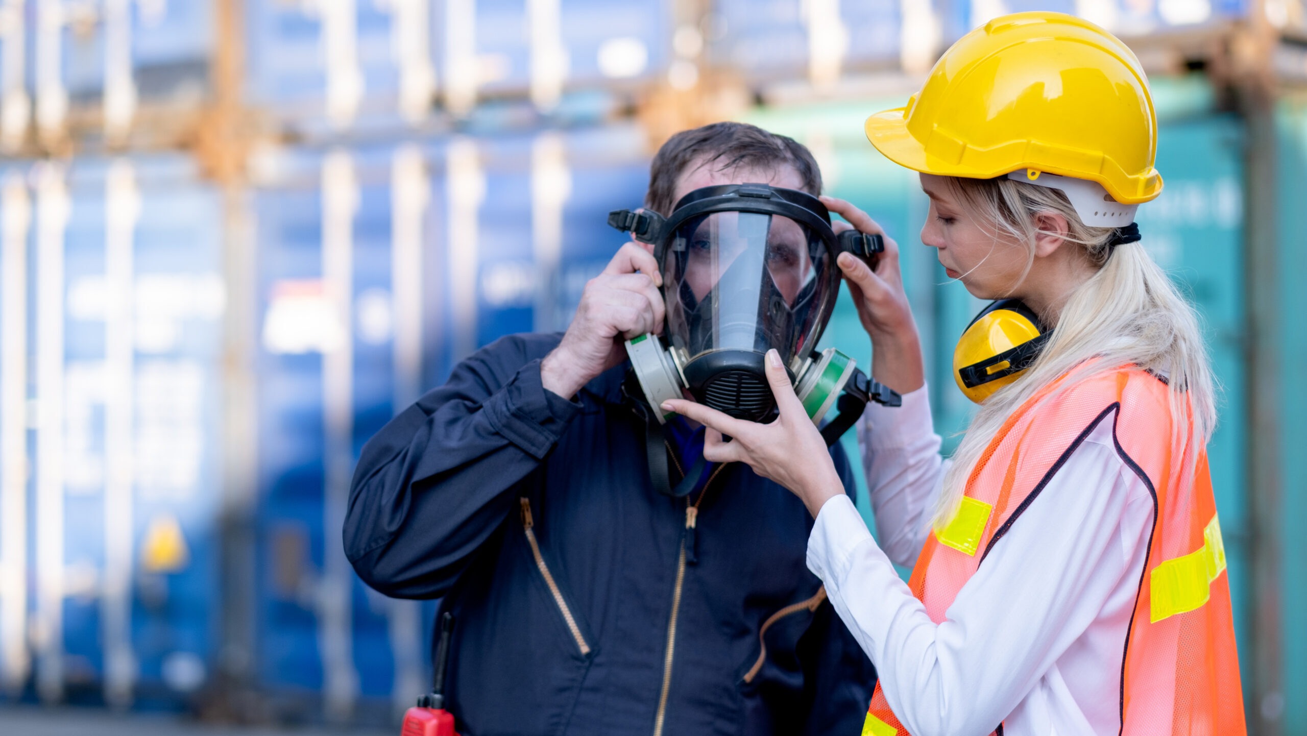 Female safety technician in a hard hat and high-visibility vest helps a worker properly fit a full-face respirator mask at an industrial site. The scene highlights a PPE hazard assessment in action, ensuring the correct selection and use of personal protective equipment for respiratory protection in hazardous work environments.