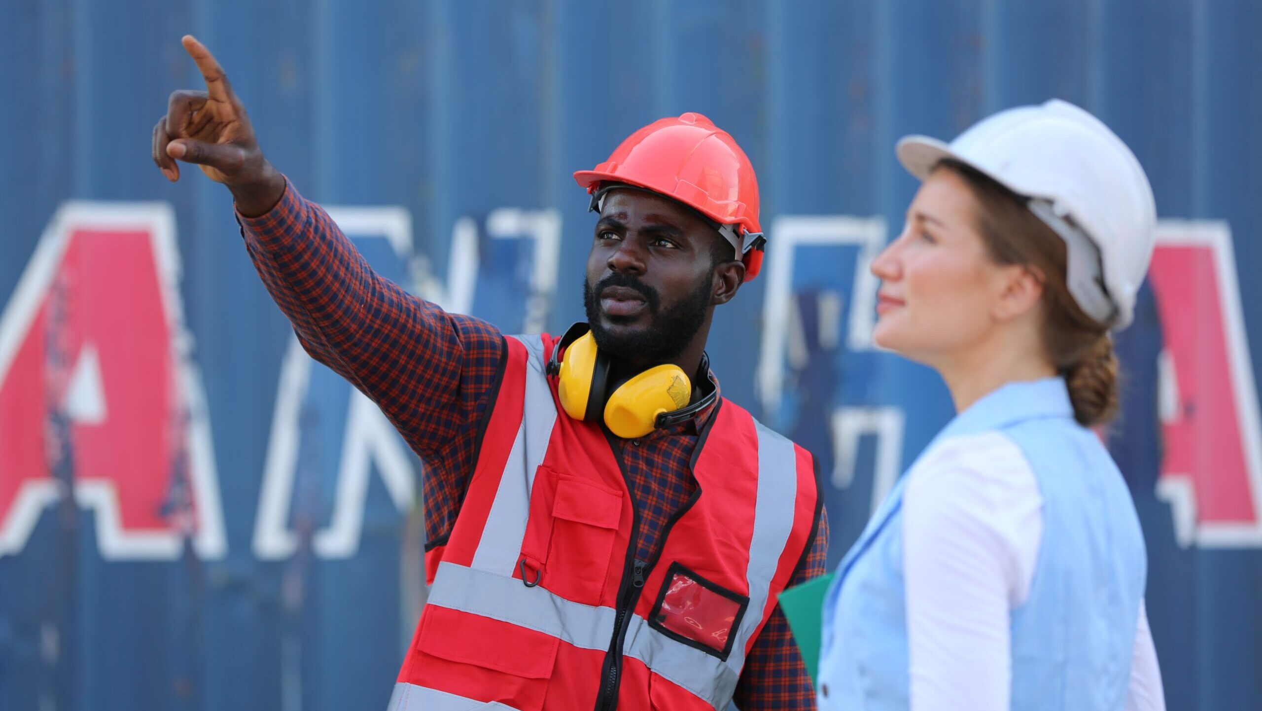 Two construction workers wearing hard hats and safety vests stand in front of a shipping container; one man points toward the distance while the woman listens attentively. This image highlights fall safety tips such as maintaining clear communication, wearing high-visibility gear, and using proper protective equipment when working outdoors in changing seasonal conditions.