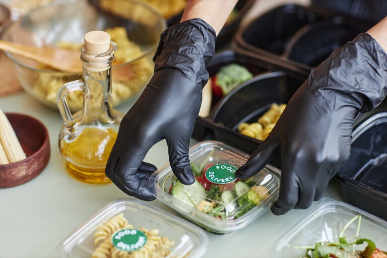 Close-up of a food service worker wearing black gloves sealing a container labeled 'Food Delivery' with fresh vegetables inside. Surrounding the container are pasta dishes and ingredients like olive oil. The image represents an allergen risk assessment in progress, highlighting safe food handling practices to prevent cross-contamination and ensure allergen labeling compliance