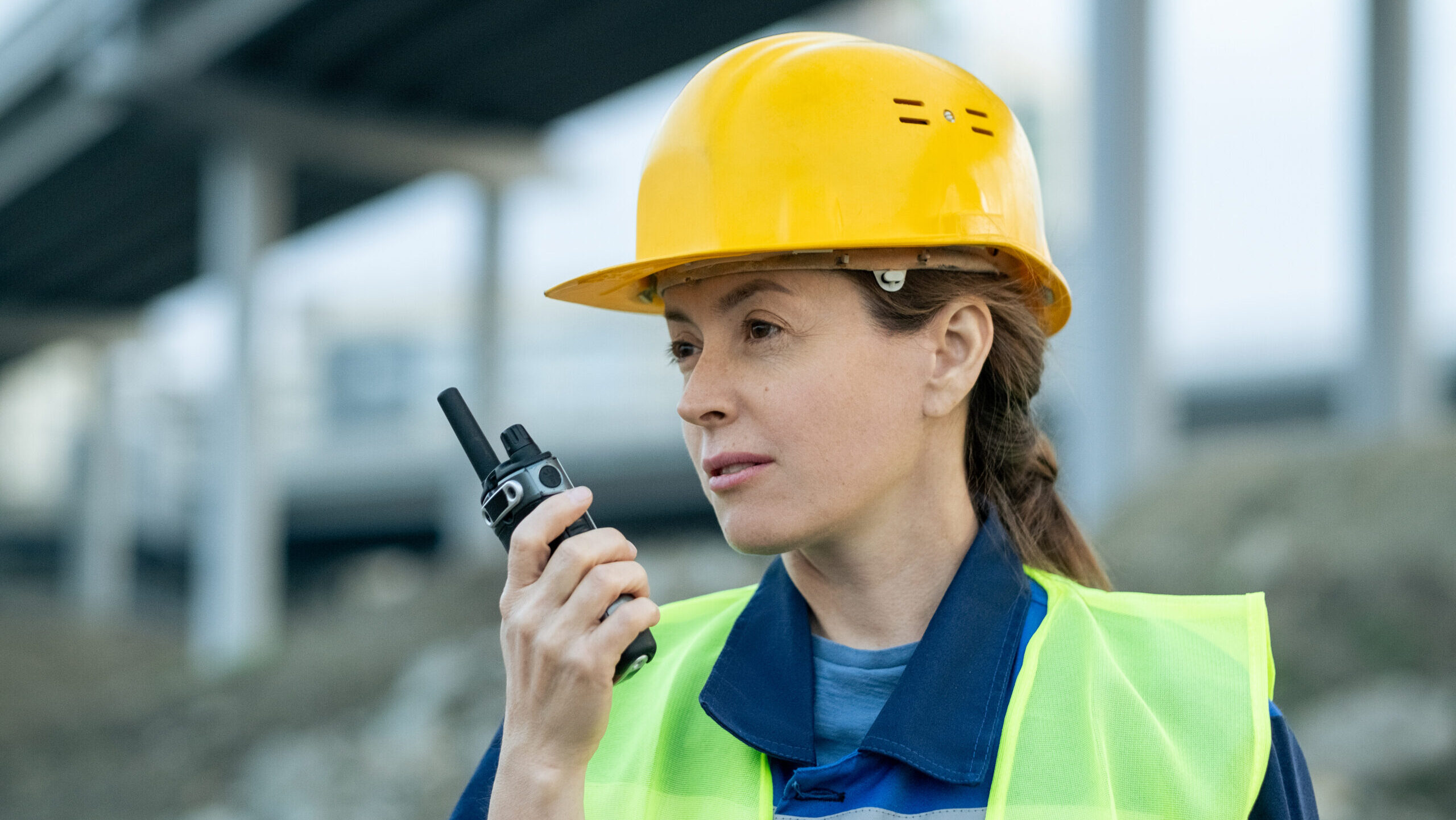 Female safety inspector wearing a yellow hard hat and reflective vest uses a walkie-talkie at a construction site—demonstrating proactive communication as part of implementing a Behavior-Based Safety (BBS) program
