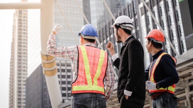Three professionals—two in construction gear and one in business attire—inspect a high-rise building site, representing companies that use contractors for urban development projects.
