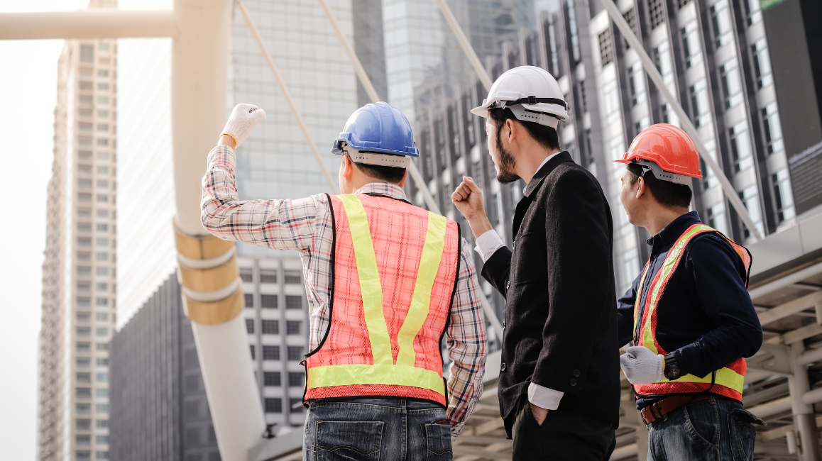 Three professionals—two in construction gear and one in business attire—inspect a high-rise building site, representing companies that use contractors for urban development projects.