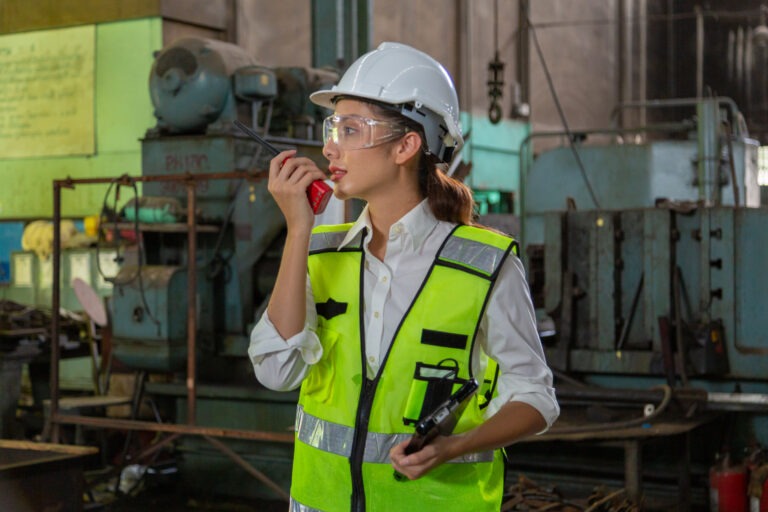 Female engineer in a high-visibility vest and hard hat uses a two-way radio while standing in an industrial facility filled with machinery. She appears to be coordinating or monitoring operations, representing a key role during benchmark events such as audits, inspections, or major process reviews in manufacturing environments.