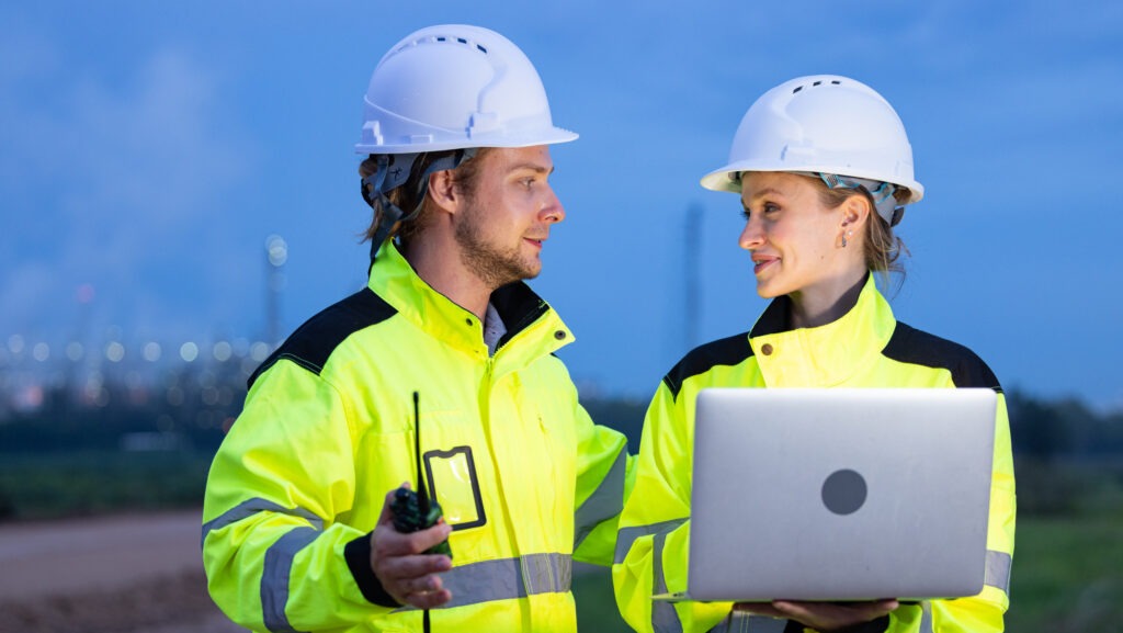 Two safety professionals in high-visibility jackets and hard hats discussing EHS tasks on-site, using a laptop and walkie-talkie—illustrating how EHS software can help manage your workload efficiently.