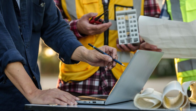 Group of engineers at a construction site reviewing building plans and data on a laptop, with one person holding a miniature building model. Rolled-up blueprints and radios are also visible. This scene represents change management examples in action, highlighting real-time collaboration, planning adjustments, and coordination across teams during a project lifecycle.