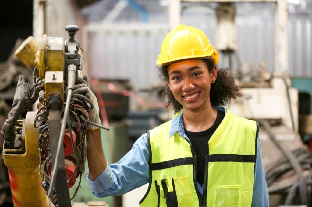 Female worker smiling, wearing a yellow hard hat and neon high visibility vest while handling a piece of heavy machinery.