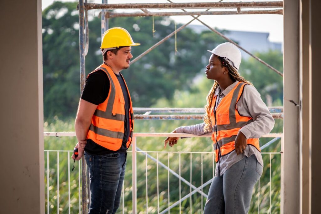 Male latino worker and black female worker in hard hats and high visibility vests having a serious discussion at a construction worksite.