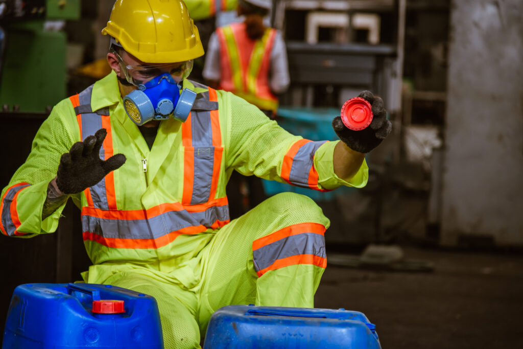 Industrial worker wearing a high-visibility suit, hard hat, gloves, and respirator while inspecting chemical containers, emphasizing chemical safety and proper PPE usage, a topic that might be covered on a JHA quiz.