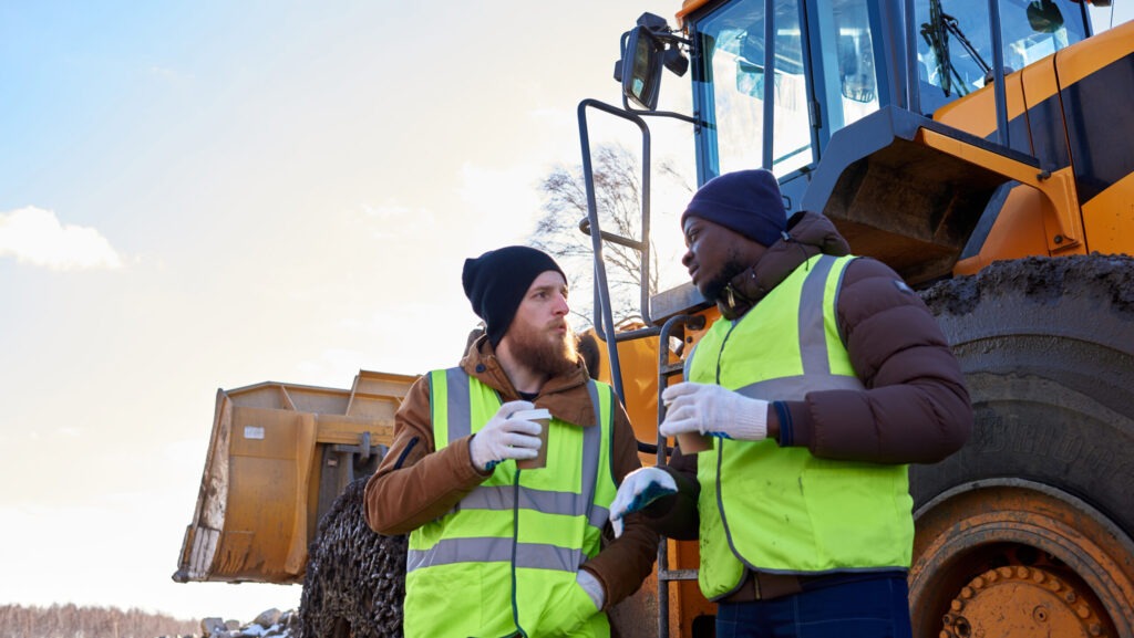 Two construction workers wearing winter jackets, gloves, beanies, and high-visibility vests stand beside heavy machinery during a break on a cold job site. They are engaged in conversation while holding hot drinks, illustrating a cold weather toolbox talk focused on safety, warmth, and preparedness in freezing outdoor conditions.