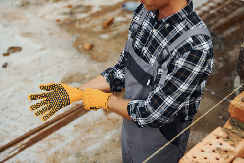 Construction worker putting on protective gloves during a hand safety toolbox talk at a job site, wearing a plaid shirt and gray overalls