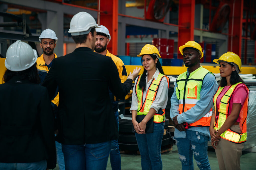 Group of industrial workers wearing hard hats and safety vests listen attentively to a supervisor during a meeting on the manufacturing floor. The scene represents a complacency toolbox talk, emphasizing the importance of staying alert, following procedures, and avoiding routine-based oversights in a high-risk work environment.