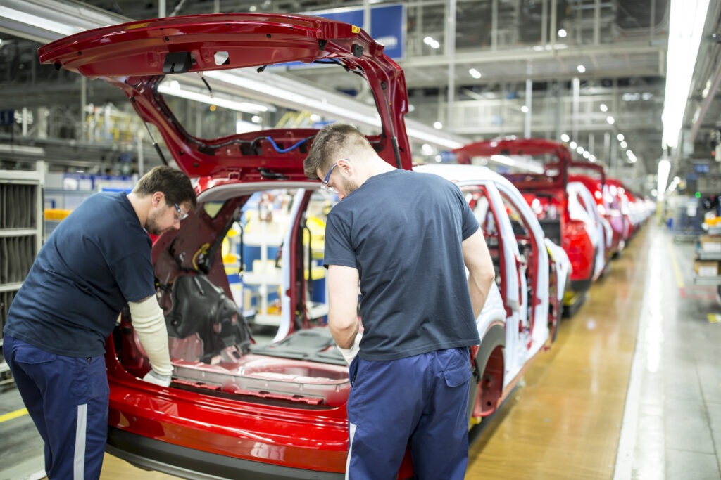 Two assembly line workers in a car manufacturing plant working on the rear section of a red vehicle, representing production downtime risks in automotive manufacturing.