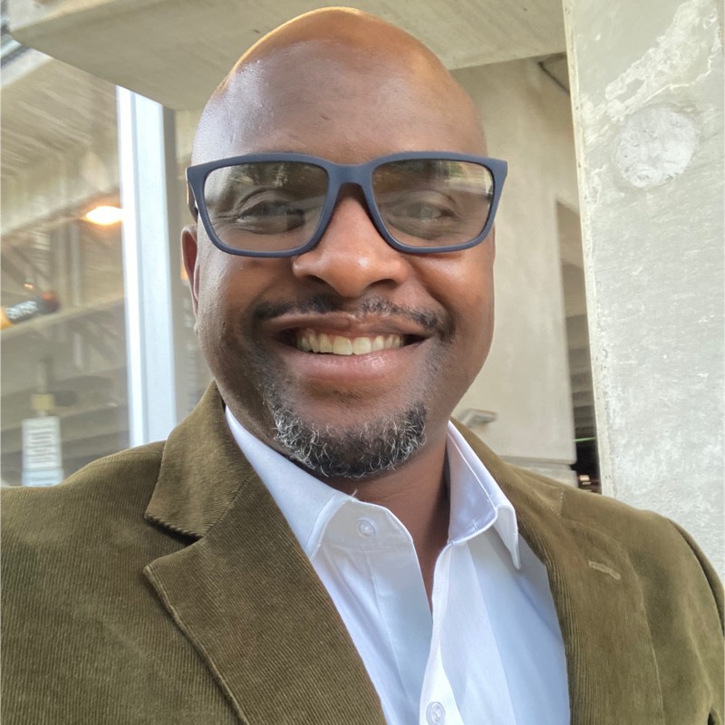 Davenj Bradley smiling while wearing glasses and a brown suit jacket with a white button down shirt as an EHS expert on risk identification and mitigation.