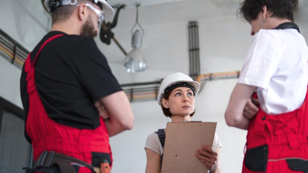 Supervisor discussing how to write a statement about an incident at work with two employees in a factory setting, using a clipboard to document incident details.