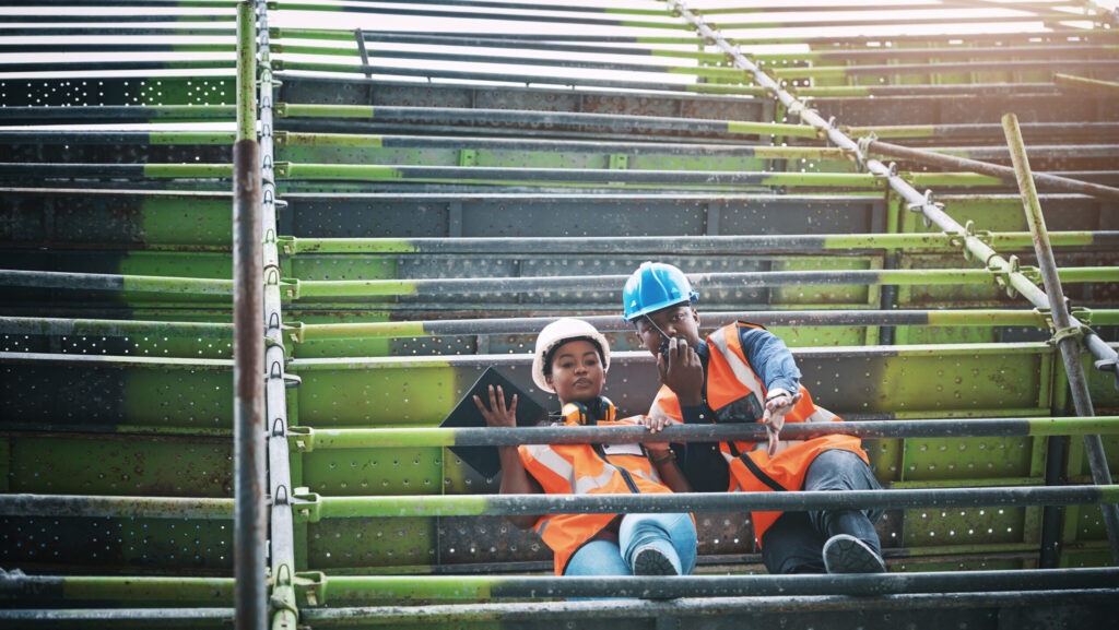 Two construction workers in safety vests and hard hats discussing plans on scaffolding, illustrating the concept of proactive risk management on a job site.