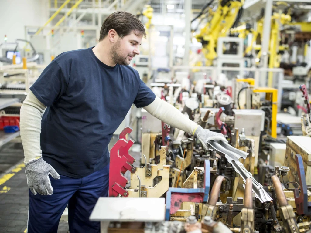 Male worker in blue pants and shirt, handling a piece of metal in an industrial setting.