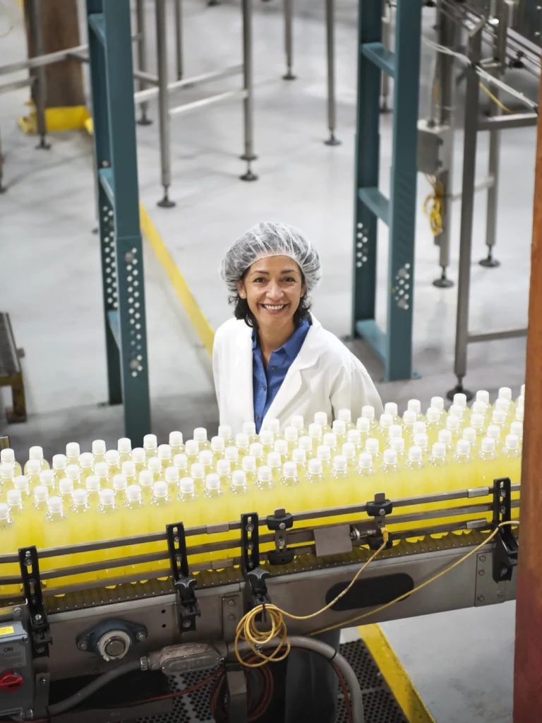 A female worker in a hair net and white coat smiling behind a beverage bottling conveyor belt.