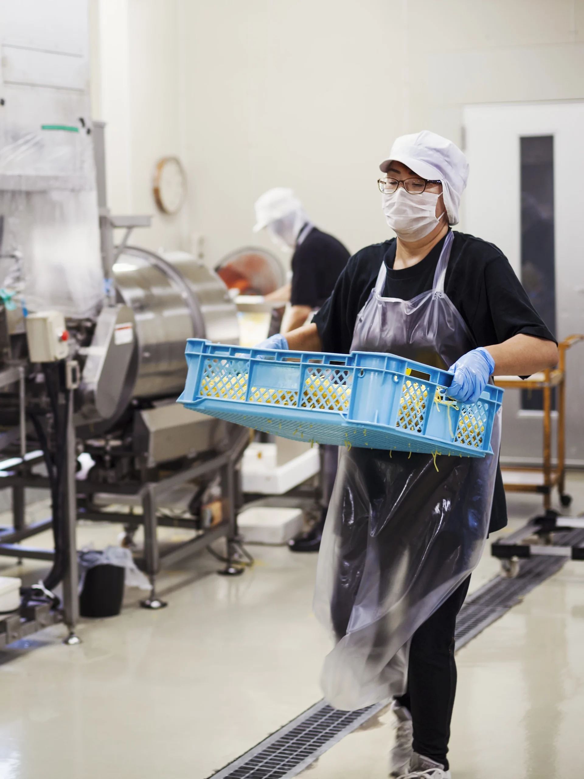 Two workers in a food production facility making soba noodles. One worker is carrying a blue crate and the other is in the background doing an audit with food and beverage health and safety software.