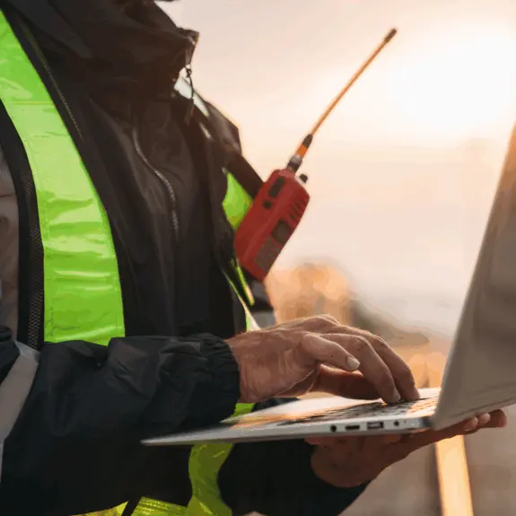 A foreman wearing a red radio and safety PPE using EHS software on a laptop.