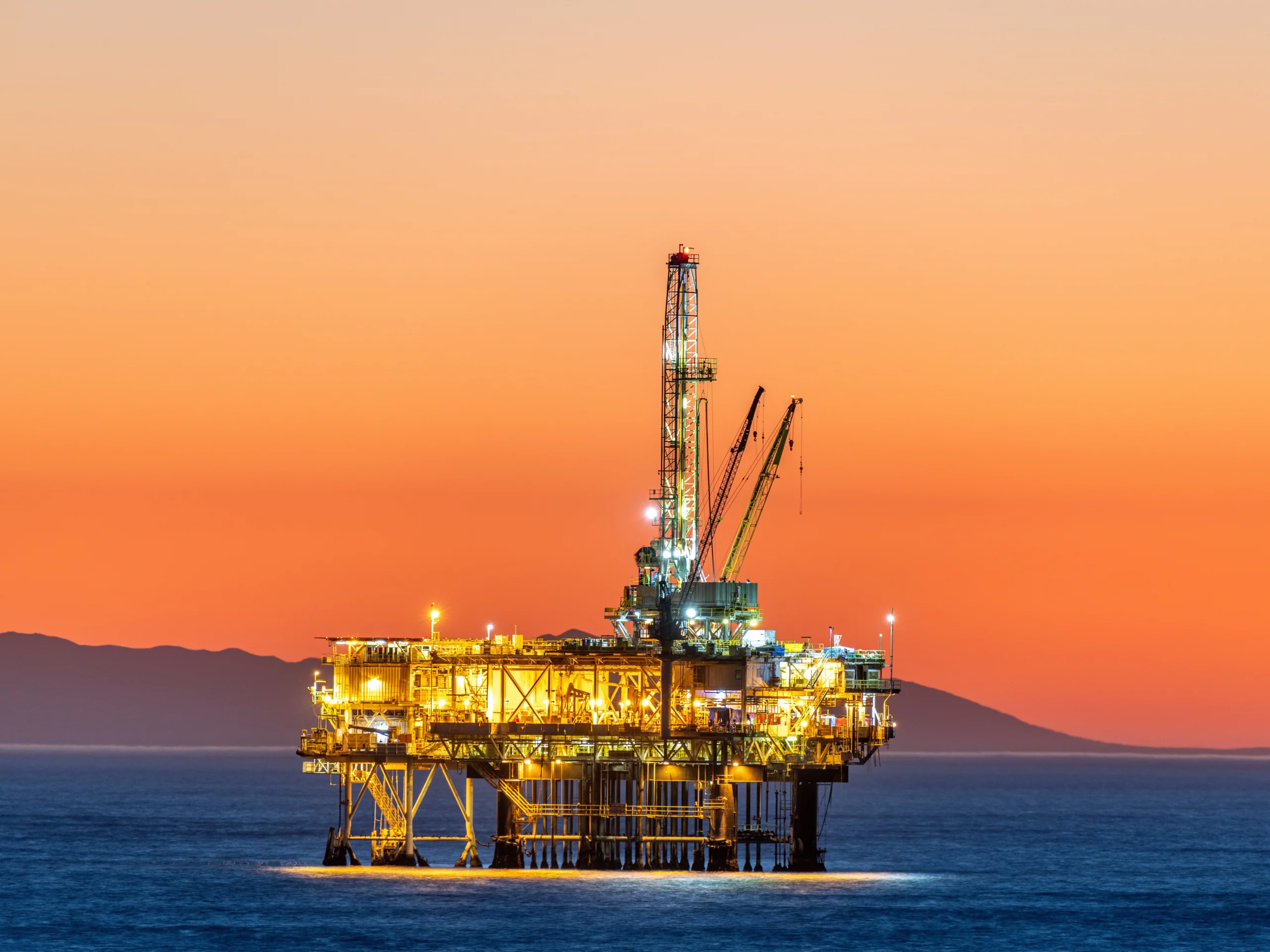 An offshore oil and gas rig in a big body of water with a land horizon behind it. The sky is orange because of the sunset.