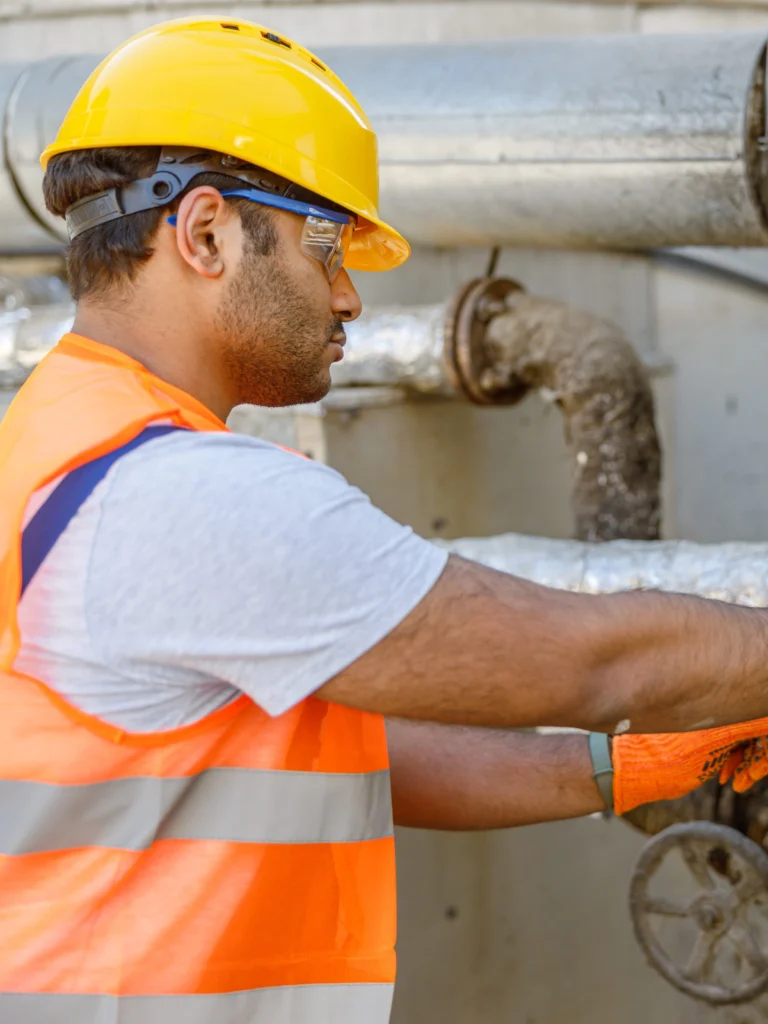A male oil rig worker in a yellow hard hat, white tshirt, orange high visibility vest, and orange gloves turns a valve.