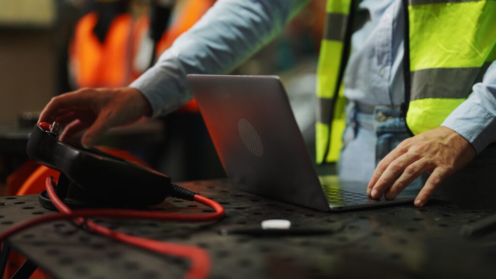 Close-up of a safety engineer in a high-visibility vest using a laptop and monitoring equipment in an industrial setting, illustrating how technology supports smarter safety decisions and improves workplace safety management.