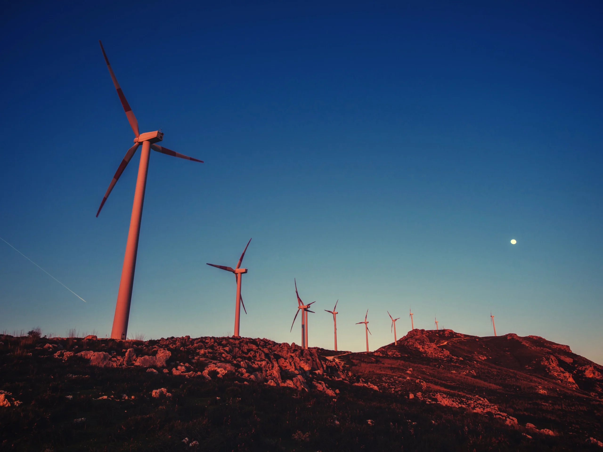 Renewable energy wind turbines in a rocky landscape with a dark blue sky and the sun going down.