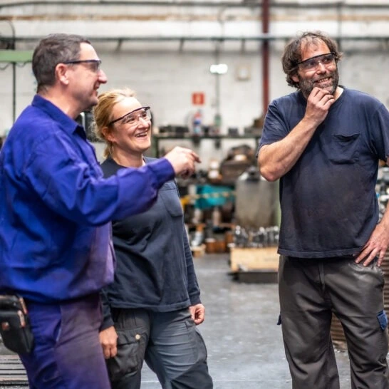 Three workers in blue work uniforms laughing in a metal fabrication shop.