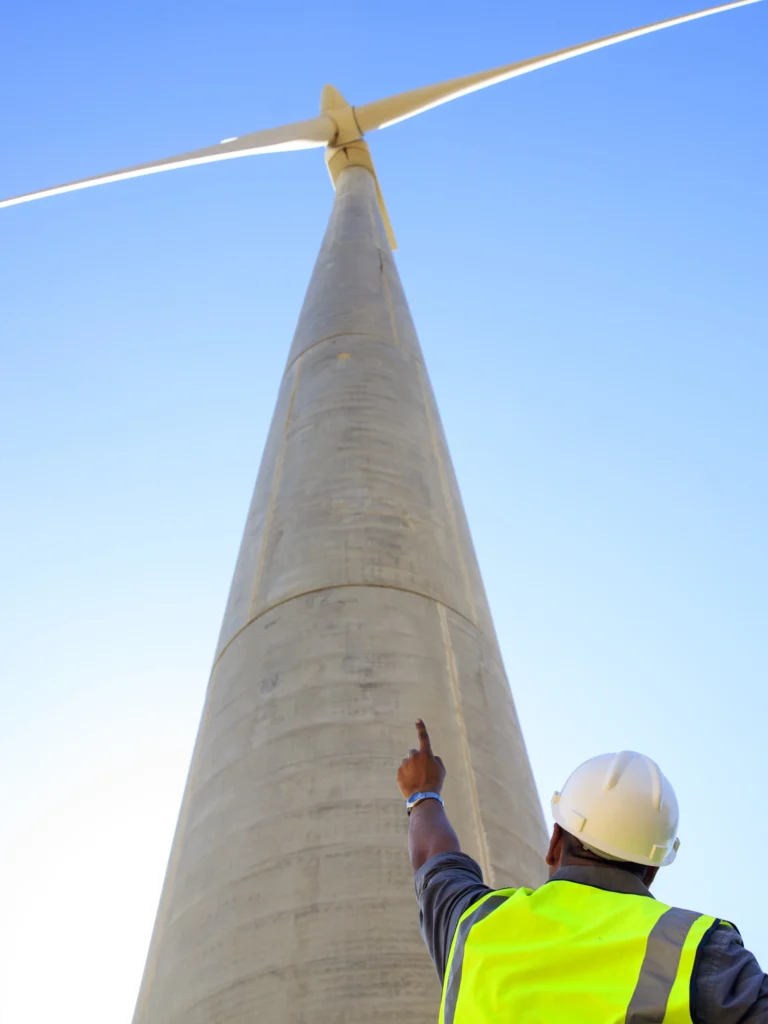 Technician in safety gear standing at the base of a wind turbine and pointing up with his left index finger.