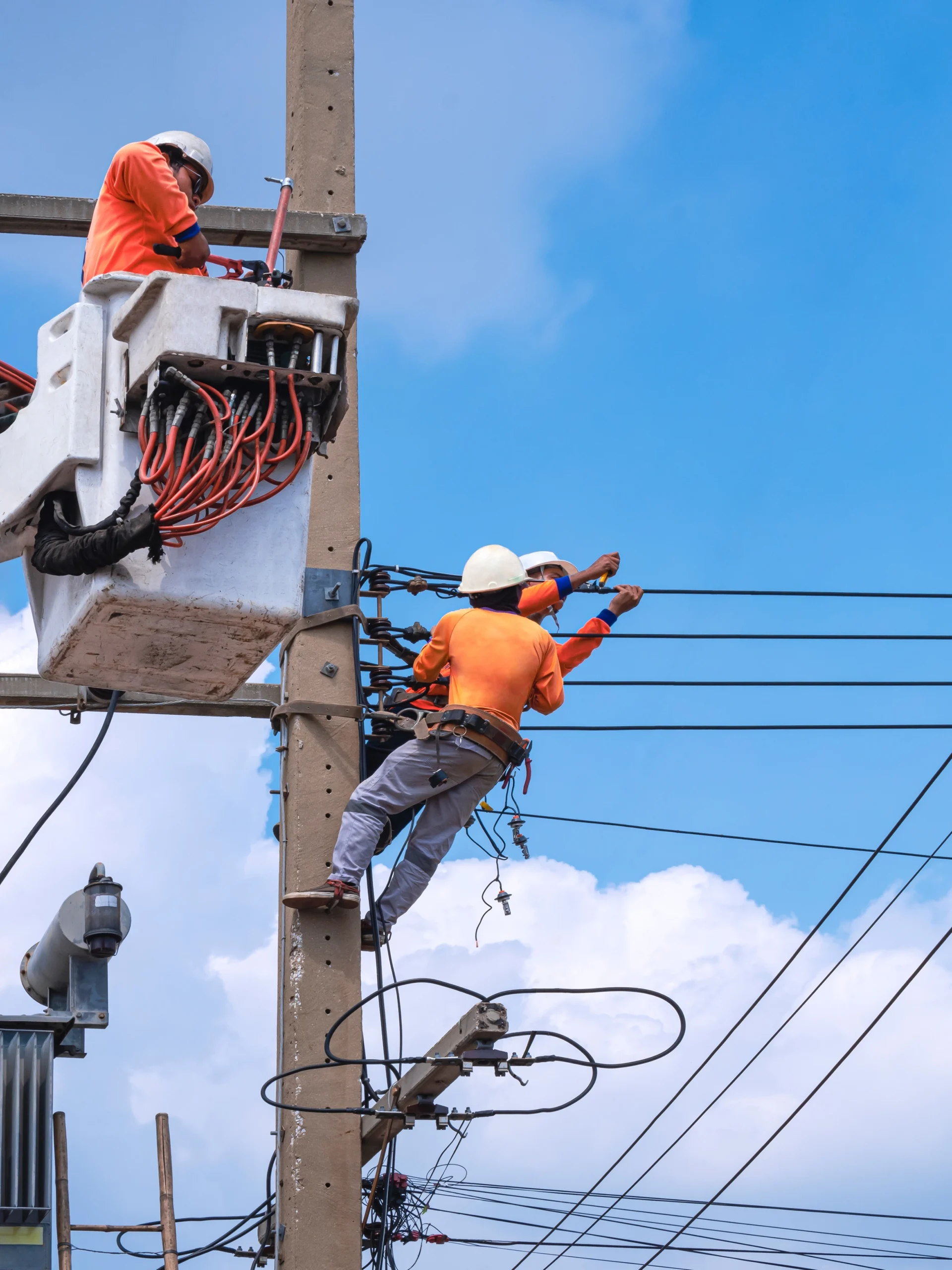 Two utilities workers in orange shirts and hard hats working on a power line.