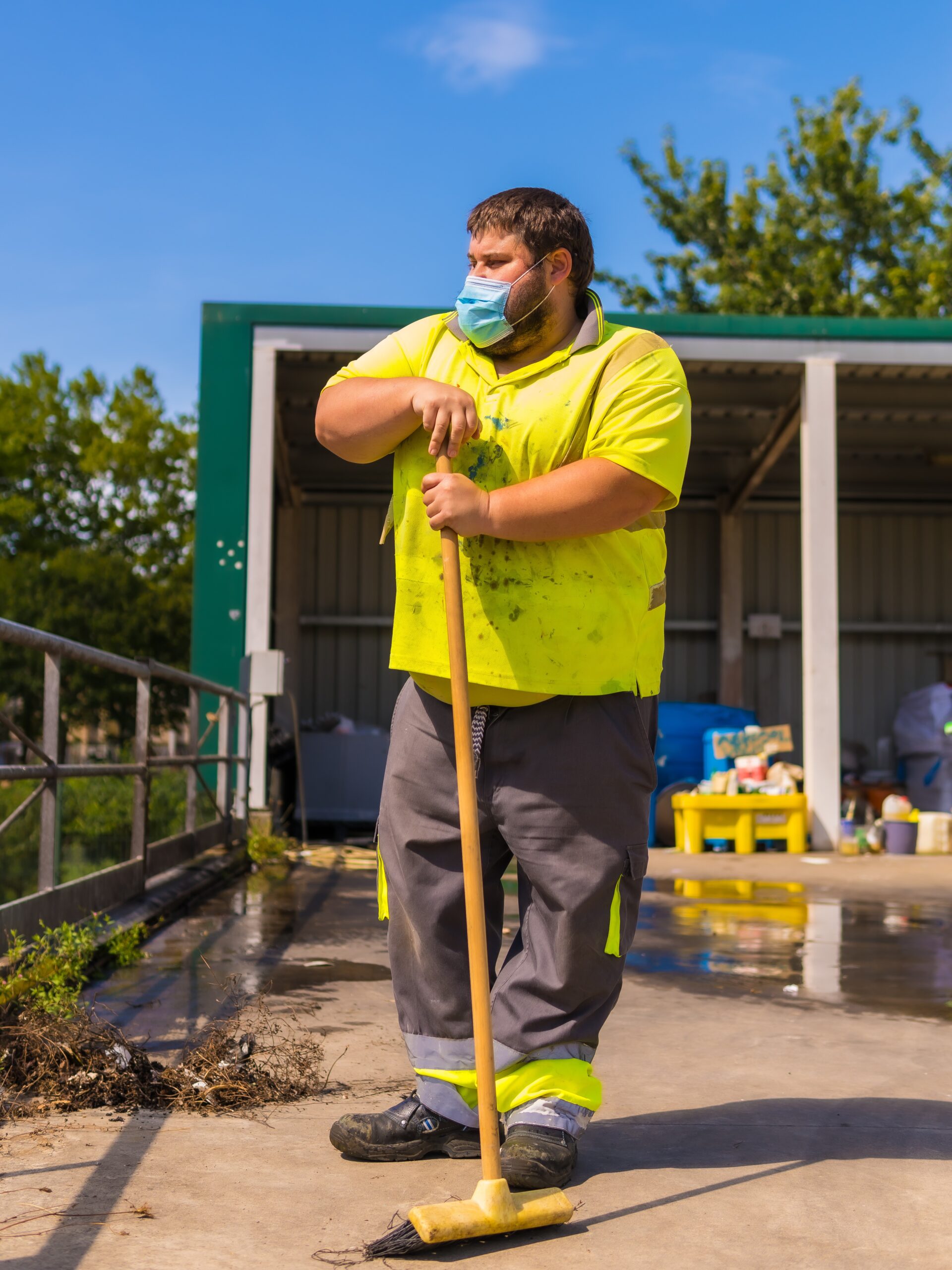 Male worker holding a push broom and standing outside in front of a municipal recycling facility.