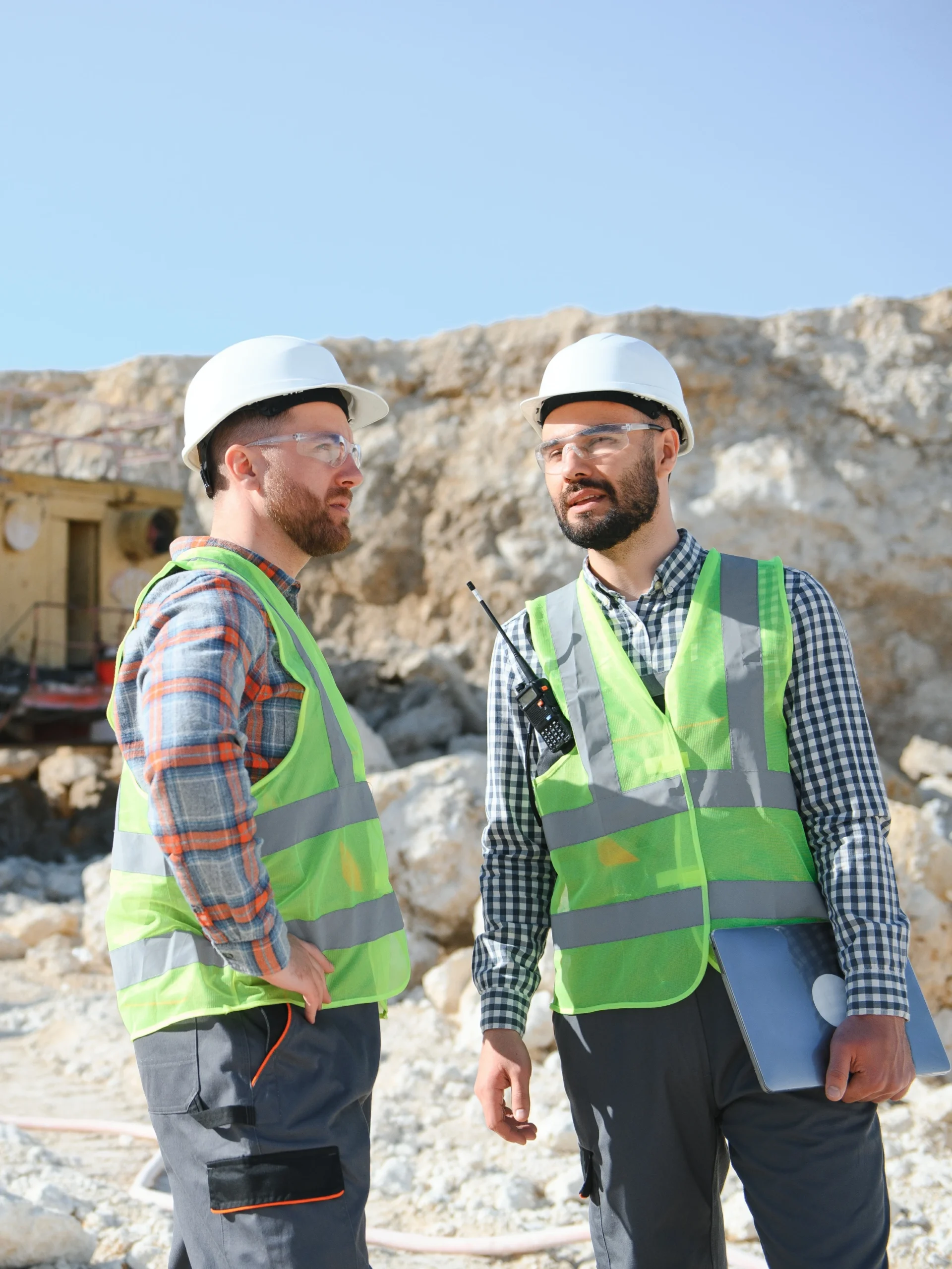 Two male workers in high visibility vests standing in a rock quarry mining location.
