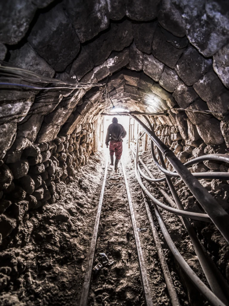 A miner walking in a silver mine with rocks and tracks on the ground.